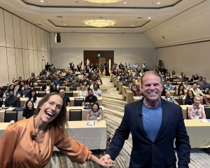 Rich and Kathy Fettke smiling standing in front of a group of RealWealth member investors at RealWealth Expo 20th Anniversary.